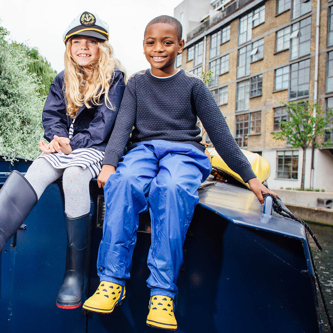 Two children wearing Originals Waterproof Trousers Blue sat on a boat. The boy is wearing blue trousers and yellow boots.